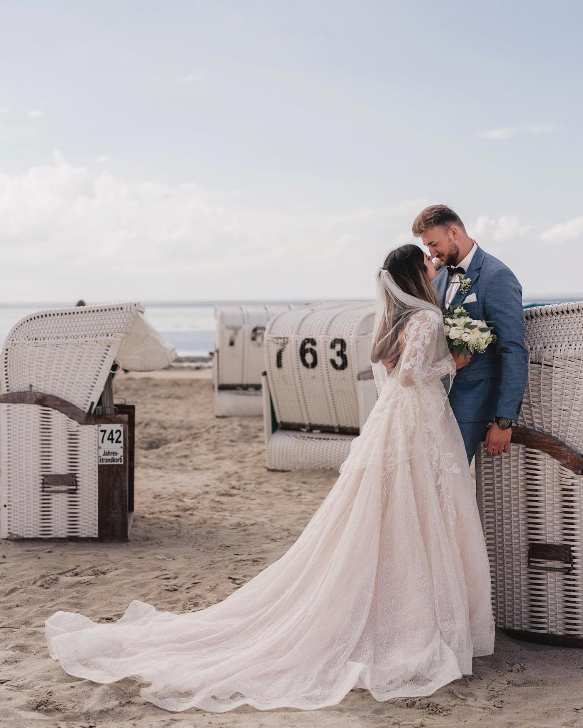 Bride and groom posing together on a sandy beach with wicker beach chairs.