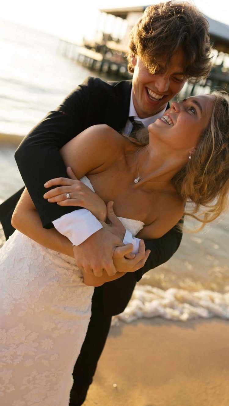 Smiling bride and groom embracing on a sandy beach with a pier behind them.