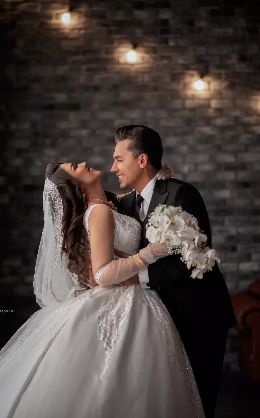 Bride and groom embracing and smiling against a dark brick wall with lights.