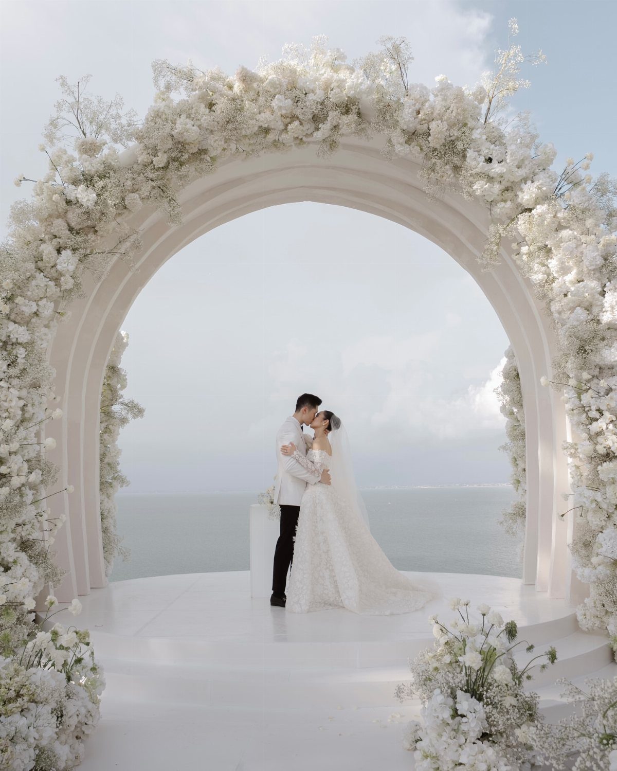 Bride and groom embracing beneath a large white floral arch by the sea.