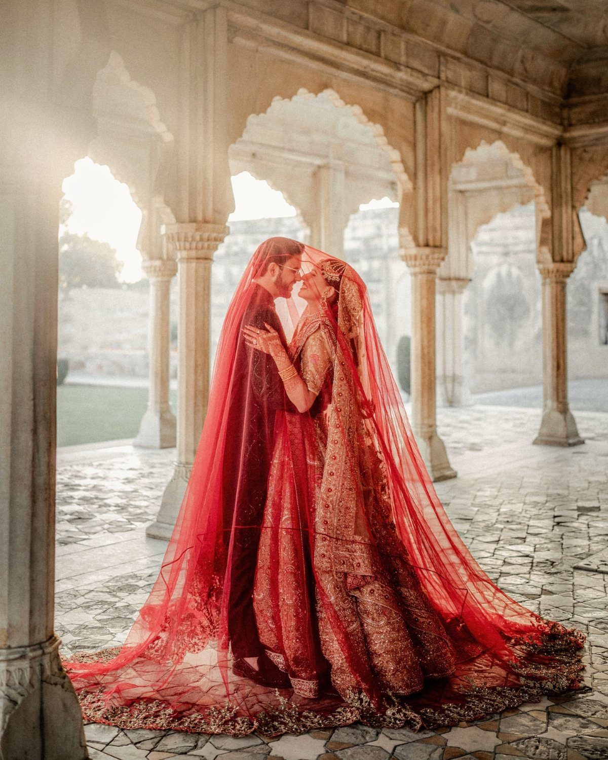 Bride and groom in red traditional Indian wedding attire posing in an ornate stone pavilion.