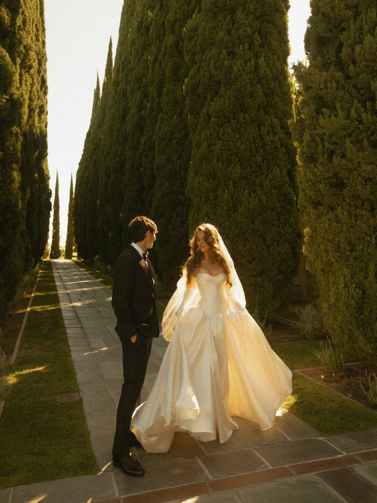 Bride and groom standing on a path lined with tall cypress trees.