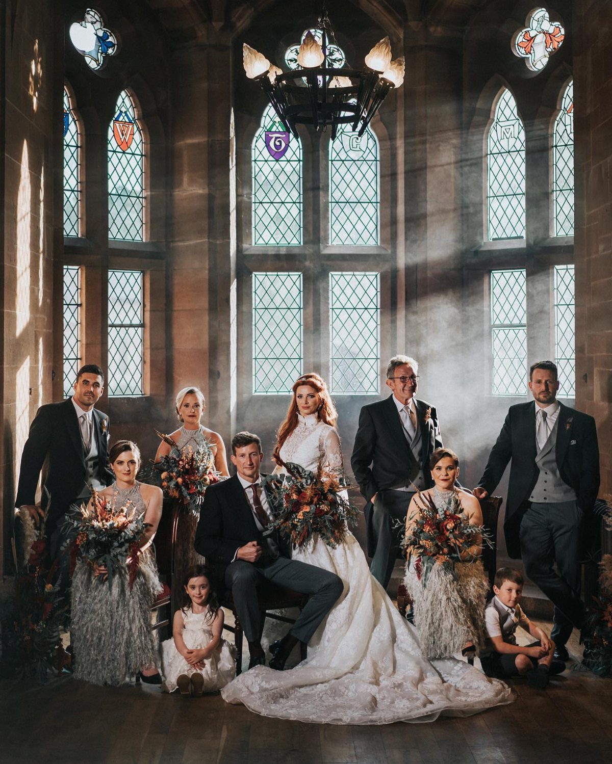 Wedding party posing in a gothic stone hall with large arched windows.