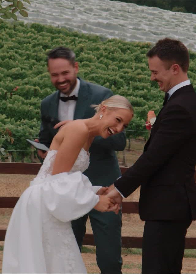 Laughing bride and groom holding hands with a smiling man in a vineyard.