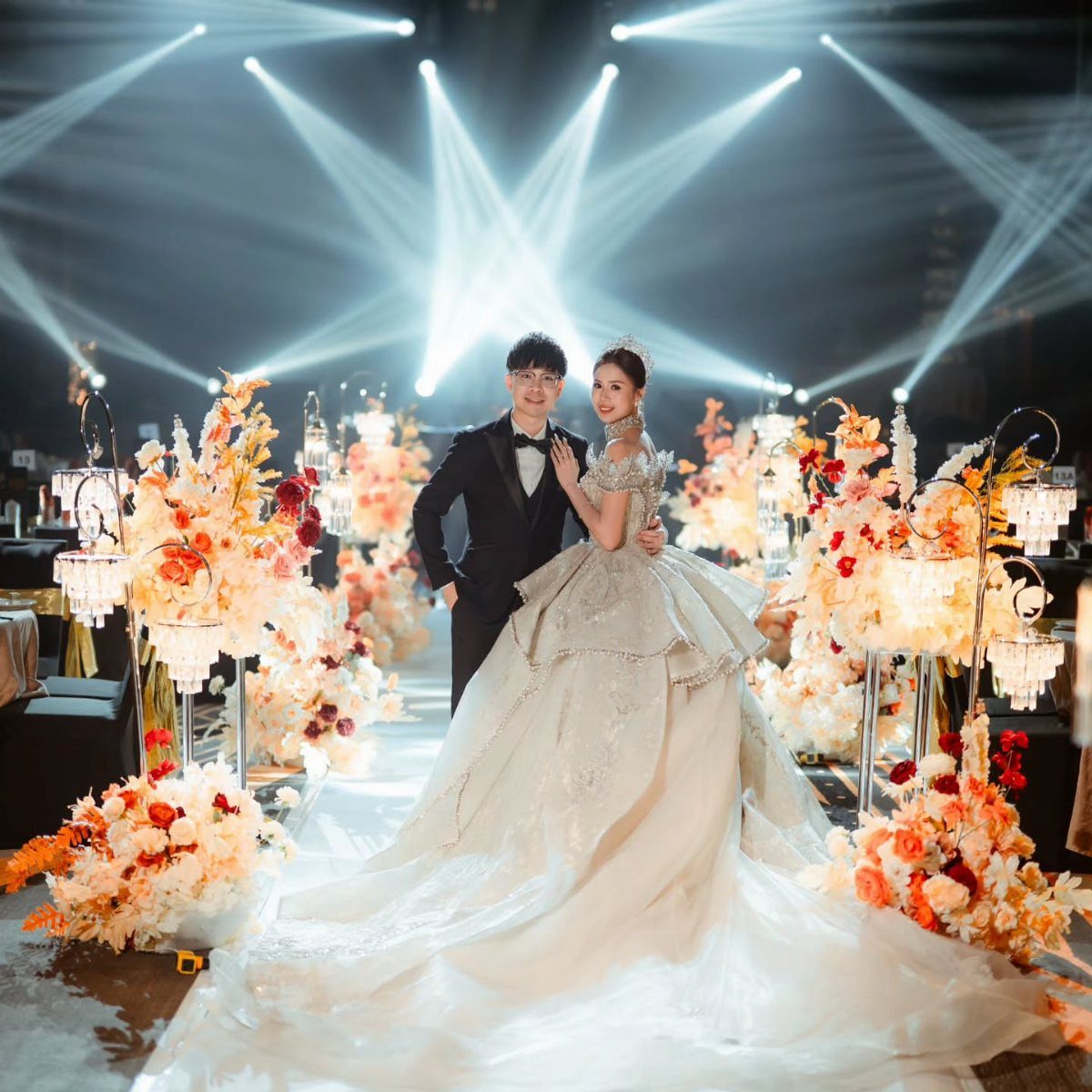 Bride and groom posing in a grand venue with floral arrangements and spotlights.