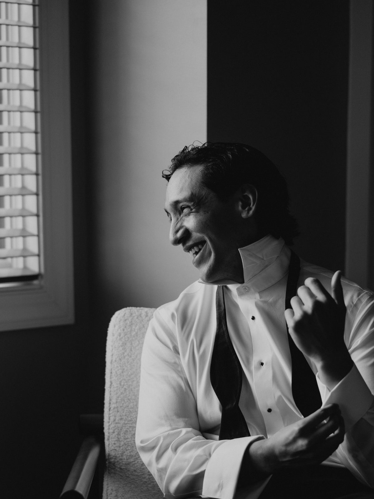 Smiling groom in a white dress shirt and tie sitting by a window.