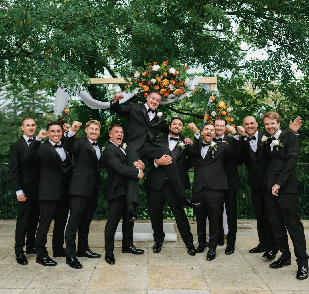 Groom in a tuxedo being lifted up by his groomsmen in front of floral arch.
