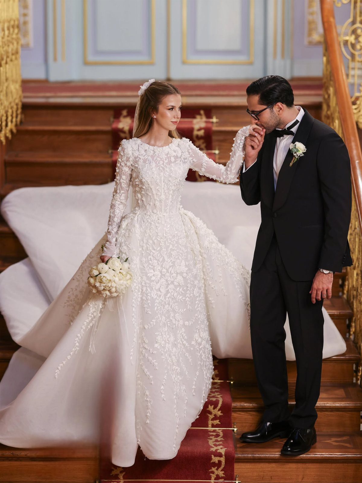 Bride in white lace gown and groom in tuxedo on wooden stairs.