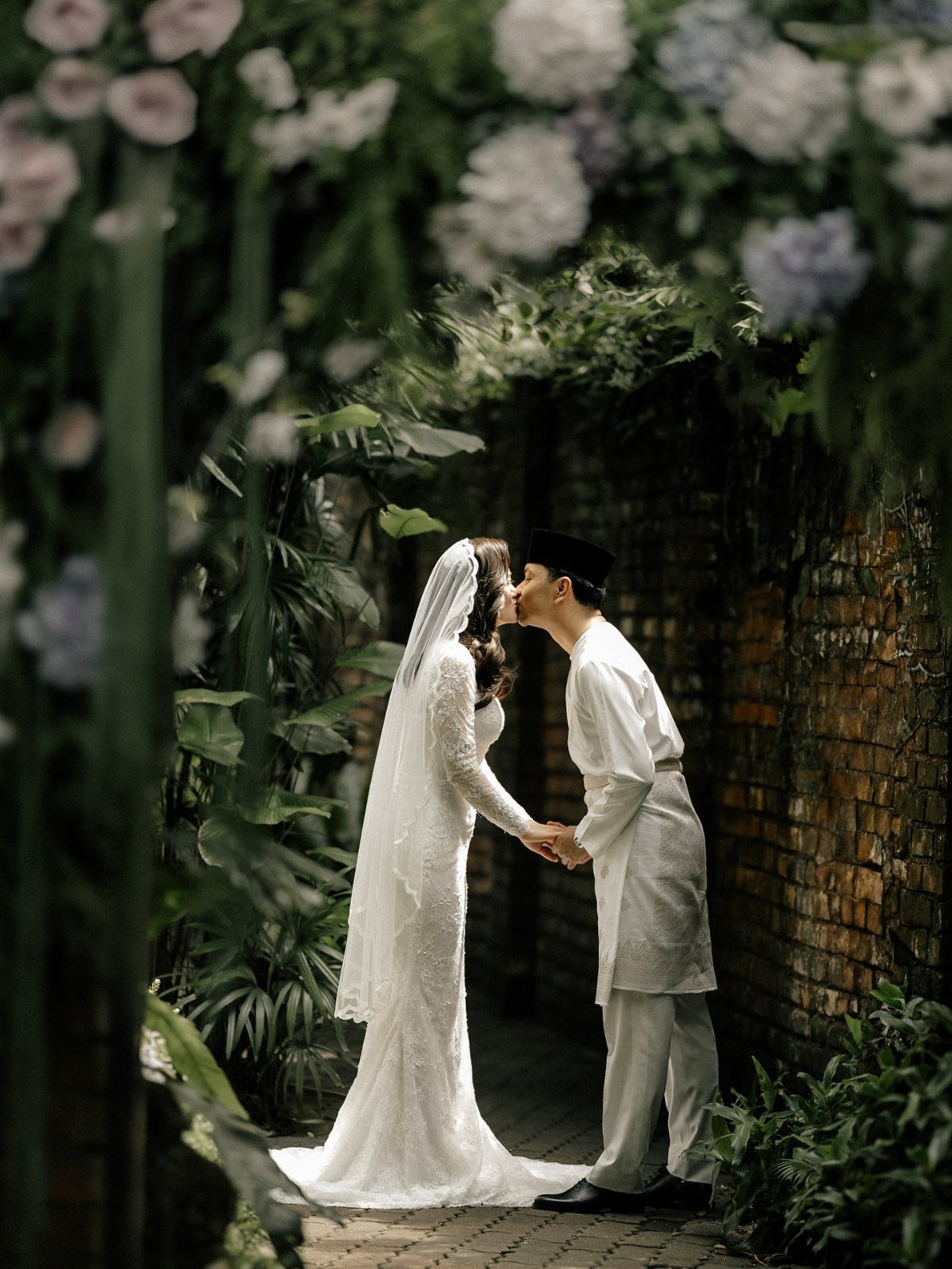 Bride and groom kissing in a lush garden path surrounded by white flowers.