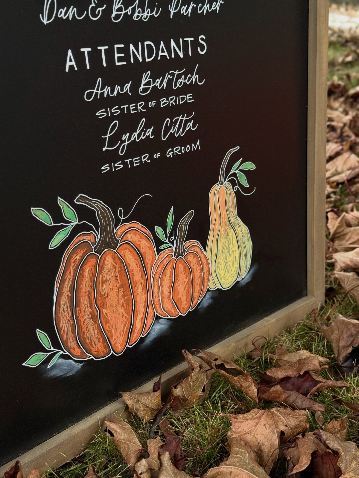 Chalkboard sign with wedding attendant names and colorful autumn pumpkin illustrations.