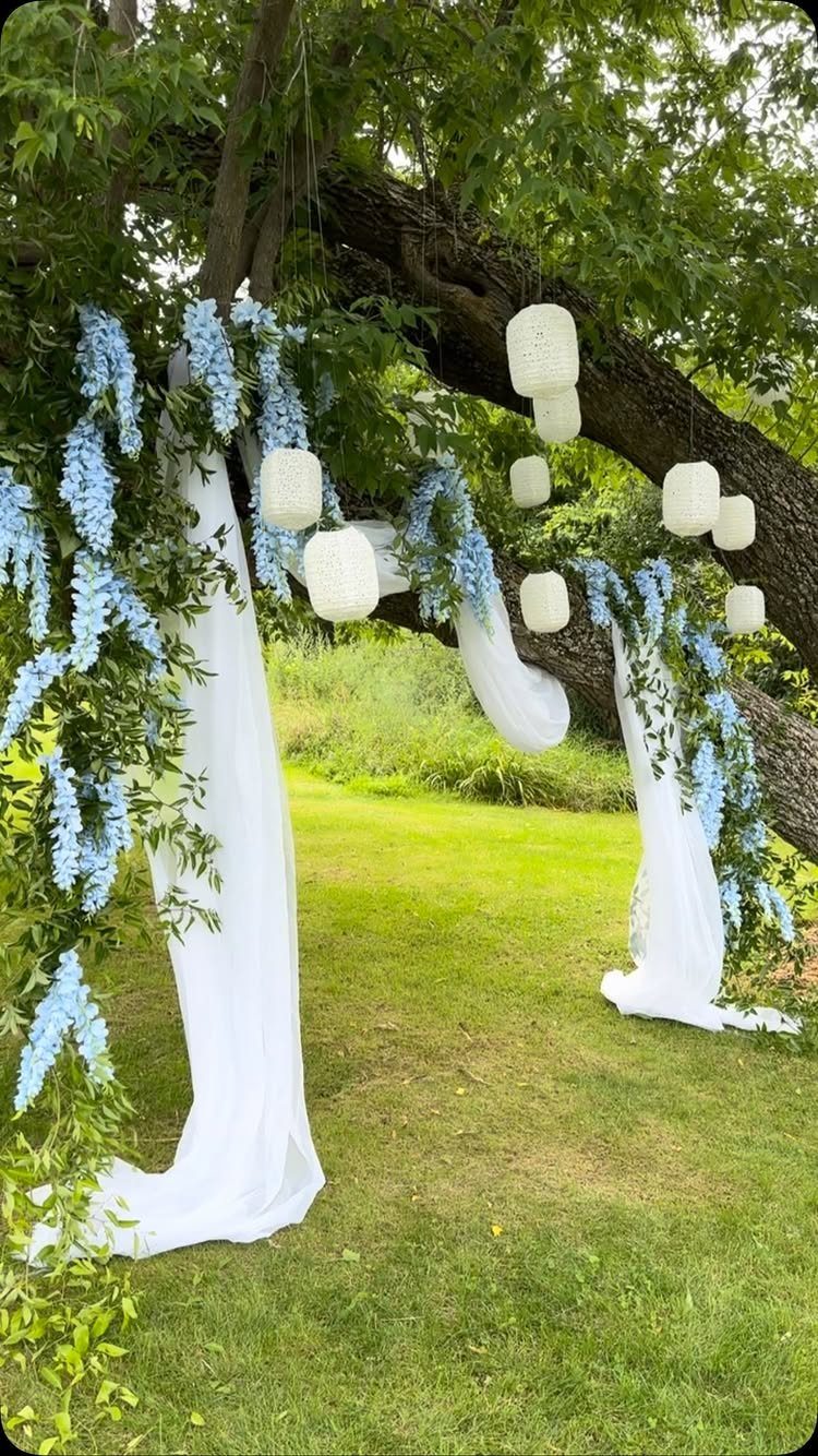 White paper lanterns and blue floral garlands hanging from a large tree branch.