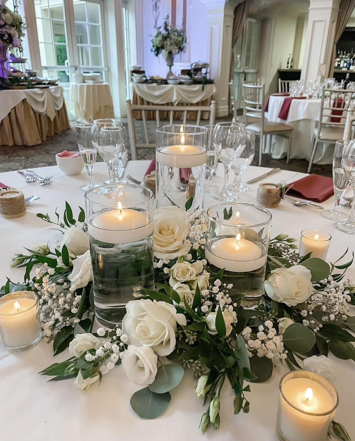 14 Candle Table Decor Ideas for Centerpieces That Feel Considered 2 Floating white votive candles in glass cylinders surrounded by white roses and greenery on a table.