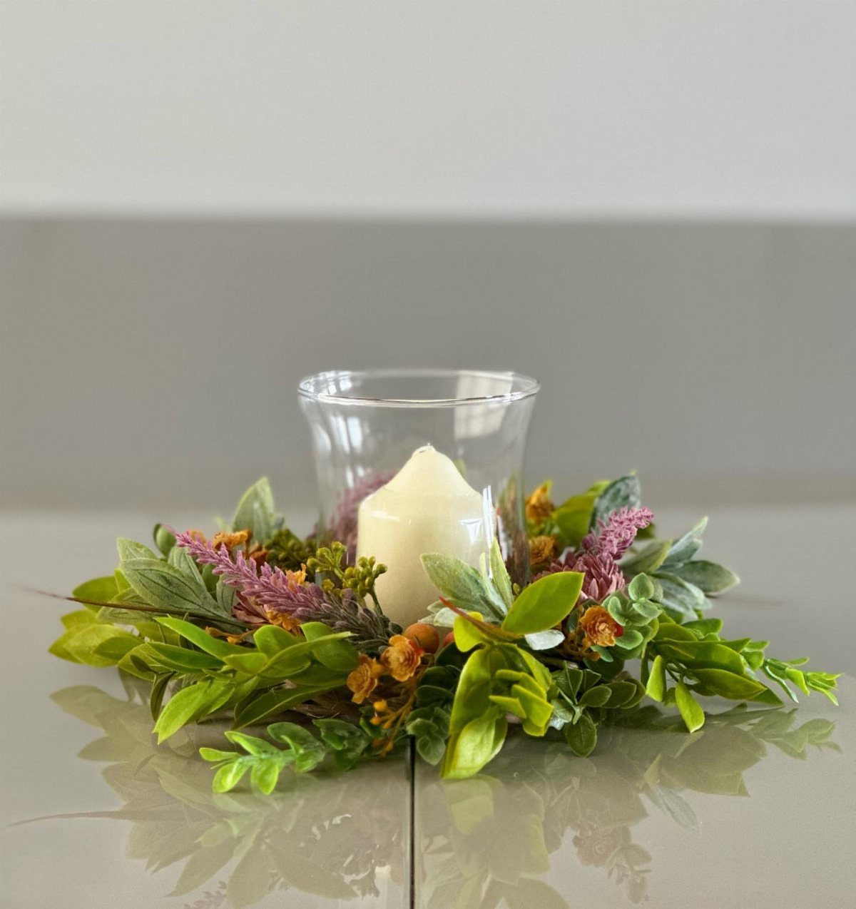 14 Candle Table Decor Ideas for Centerpieces That Feel Considered 3 A white pillar candle inside a glass hurricane holder surrounded by a green leafy wreath.
