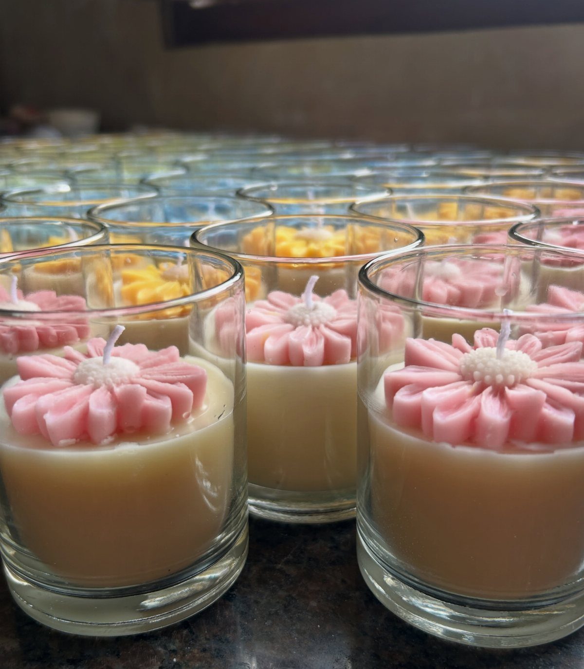 9 Candle Wedding Favors: From Beach Gel to Flower-Topped Votives 10 Small glass votive candles with pink wax flower decorations arranged in rows on a counter.