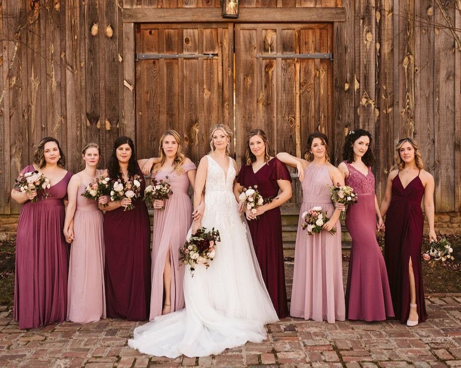21 Color Combinations for Wedding Palettes in 2026 11 Bridesmaids in burgundy and dusty rose dresses standing with a bride in front of a wooden barn.