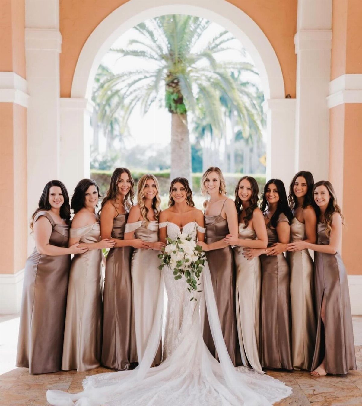 21 Color Combinations for Wedding Palettes in 2026 13 Bridesmaids in mocha and champagne satin dresses posing with a bride in white lace.