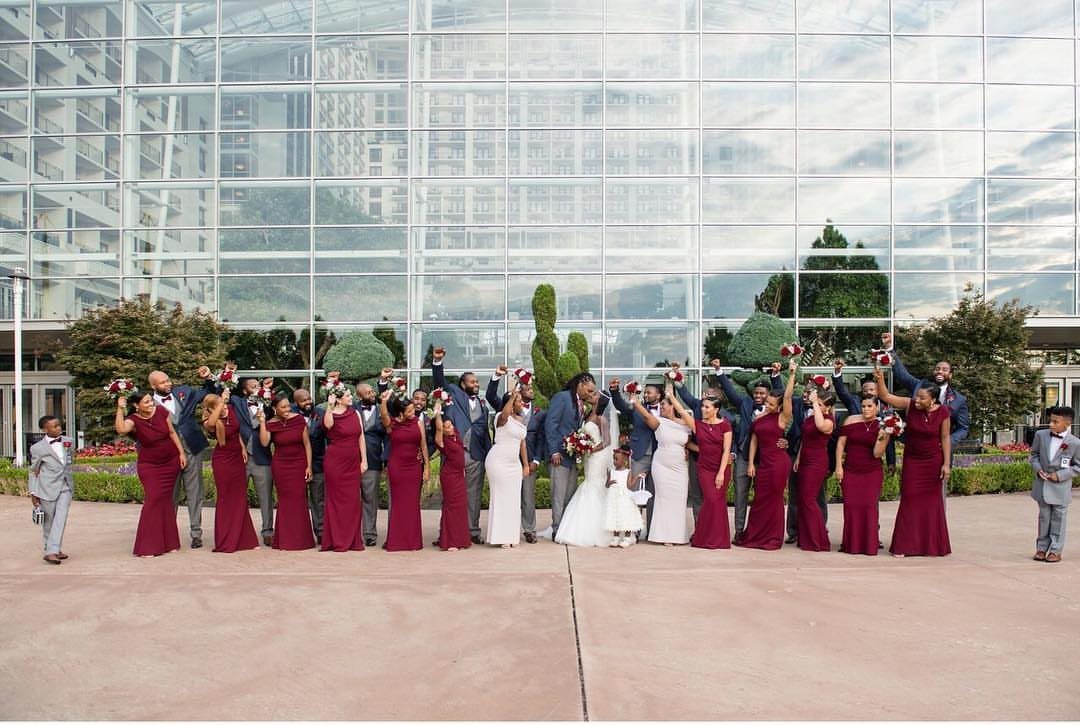 21 Color Combinations for Wedding Palettes in 2026 2 Wedding party in burgundy dresses and navy suits posing in front of a glass building.