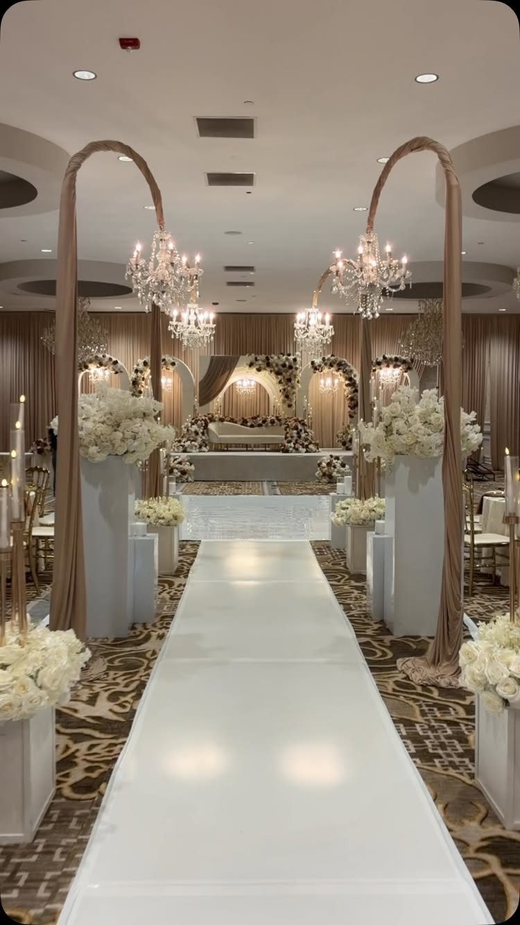 21 Color Combinations for Wedding Palettes in 2026 18 A white wedding aisle runner leads through floral arrangements and crystal chandeliers toward a decorated stage.