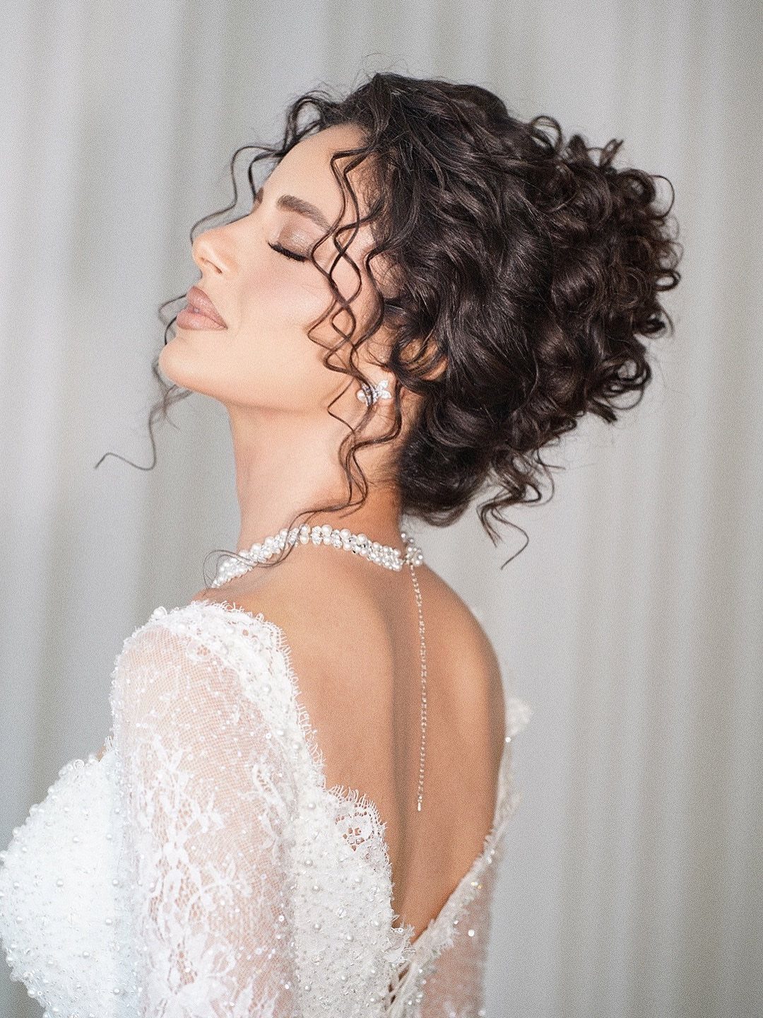 Woman with curly dark hair in an elegant updo wearing a white lace wedding dress.