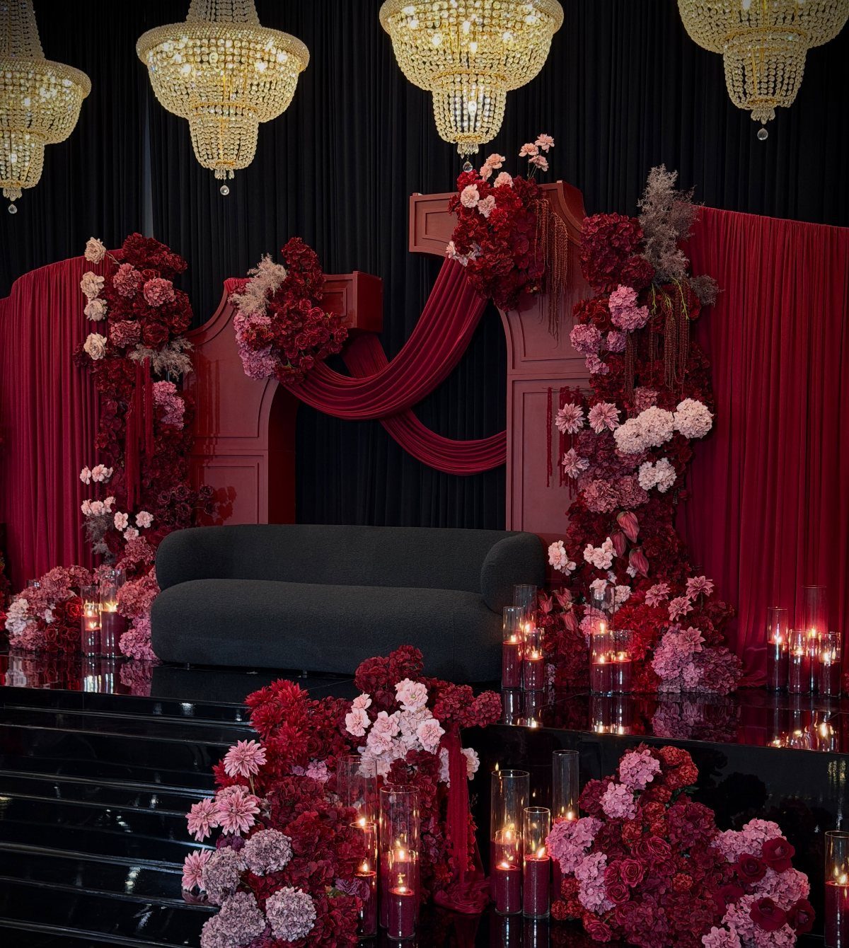 12 Dark Red Wedding Theme Ideas With Floral, Cake and Gown Picks 4 Red and burgundy floral arrangements decorating a dark wedding stage with a black sofa and chandeliers.