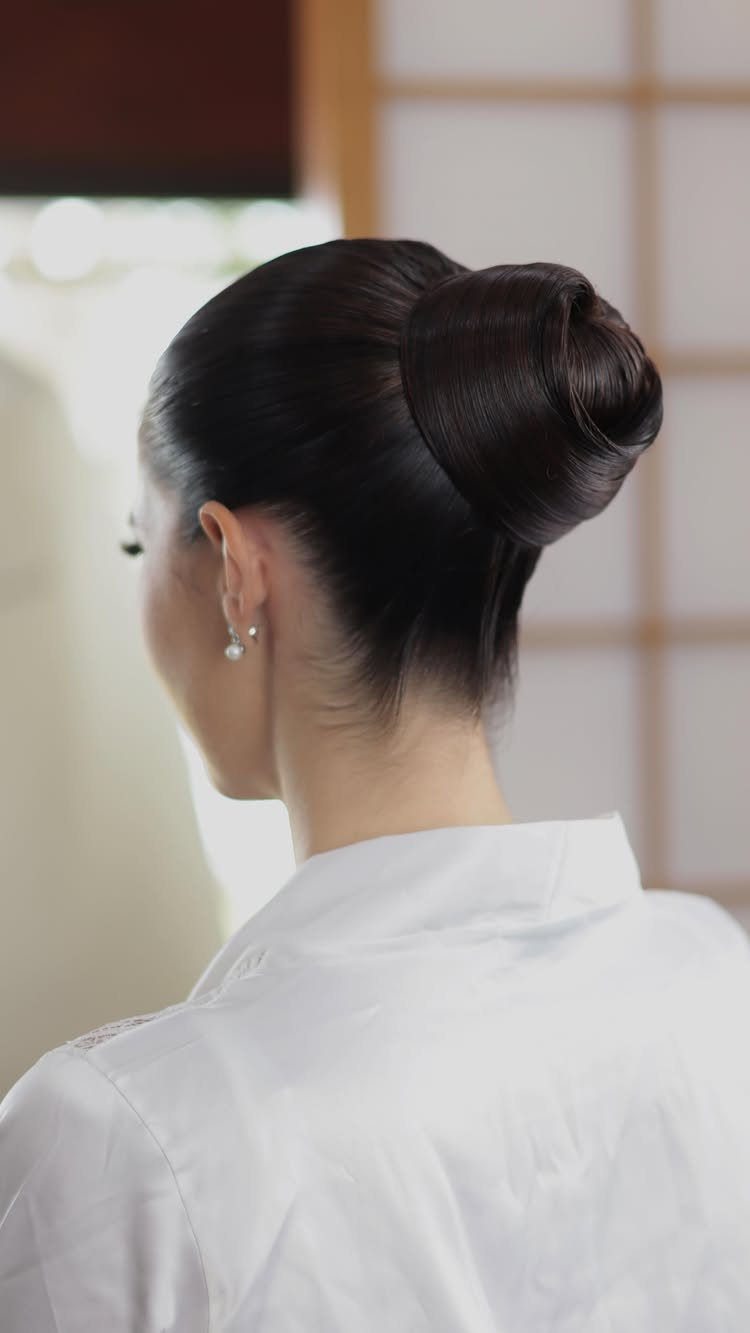 Woman with dark hair styled in a sleek, high polished topknot bun wearing a white shirt.