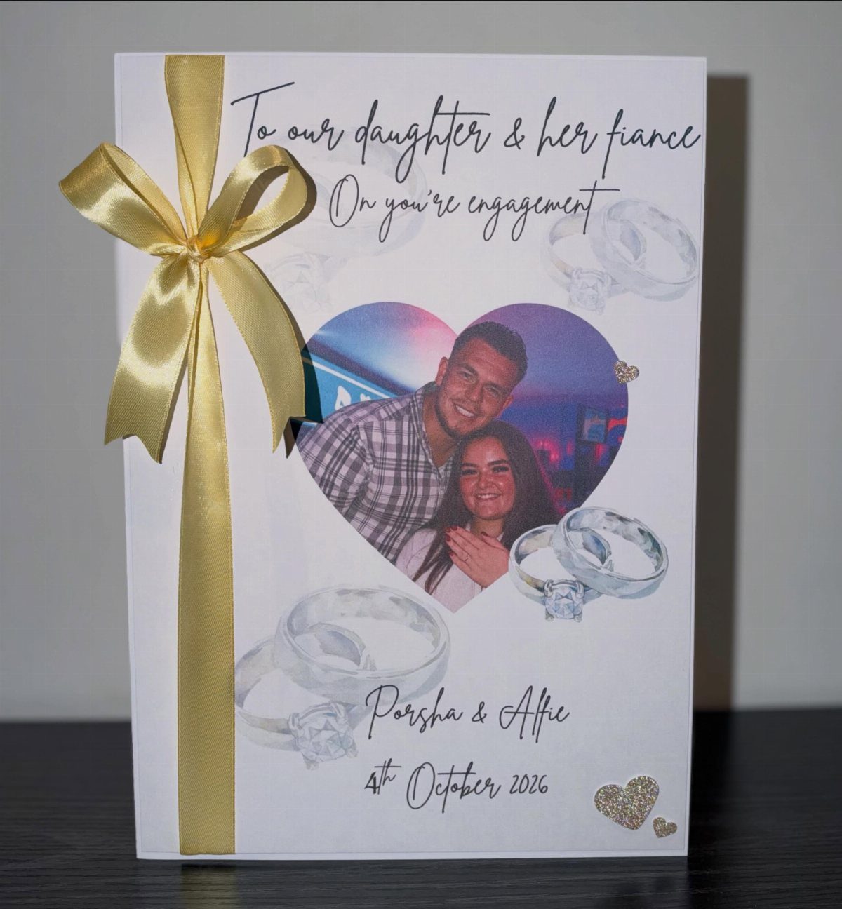 White engagement card with gold bow, heart-shaped couple photo, and diamond ring illustrations.