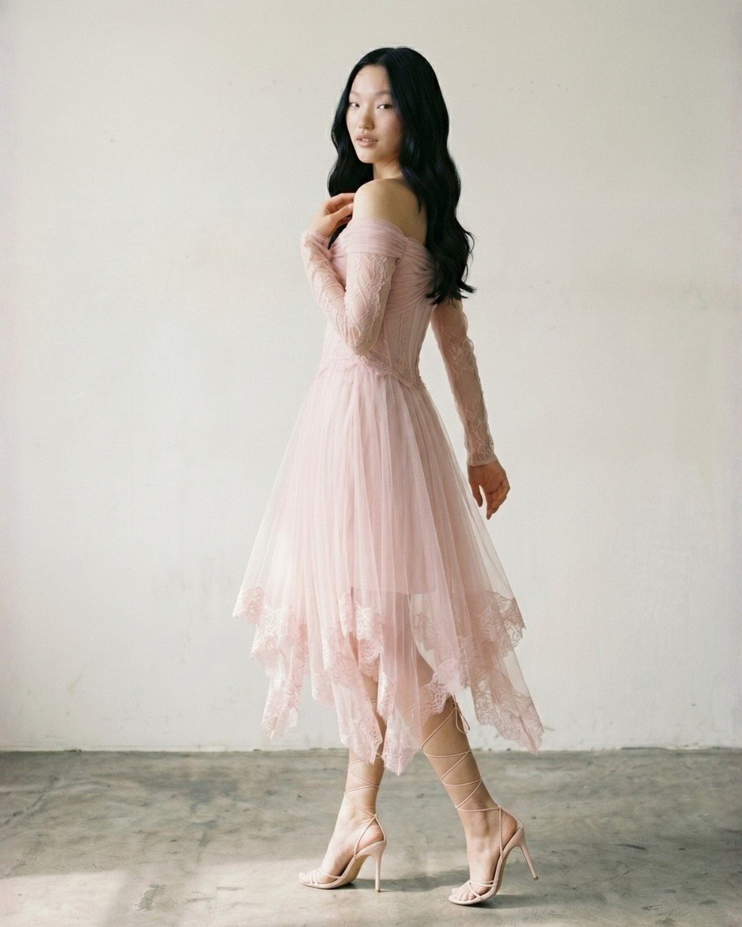 Woman in a blush pink off-the-shoulder dress with an asymmetrical lace hemline.