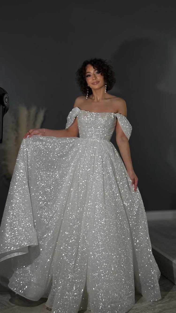 Woman in a sparkling white off-the-shoulder A-line gown posing against a grey wall.