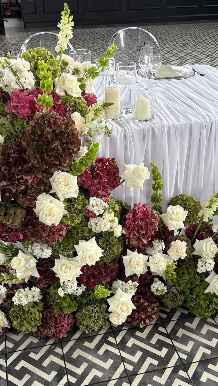 9 Fall Floral Wedding Ideas for Rich Color and Texture 6 A large cascading floral arrangement with white roses and burgundy hydrangeas draped over a white table.