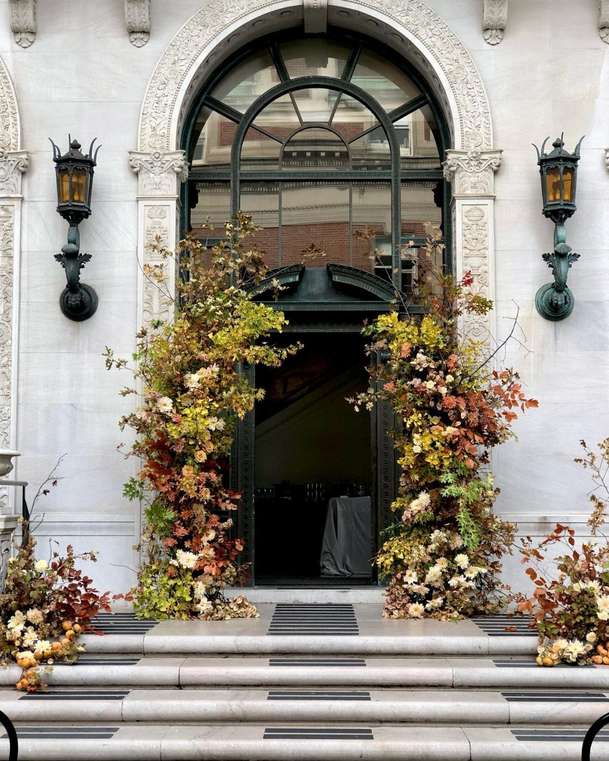 9 Fall Floral Wedding Ideas for Rich Color and Texture 9 Large autumn floral arrangements flanking a white arched stone doorway with dark metal window panes.