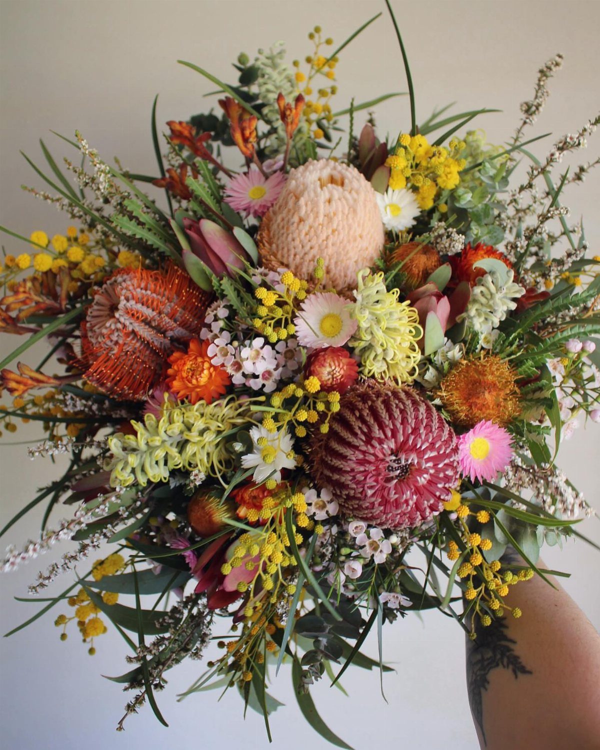 9 Fall Floral Wedding Ideas for Rich Color and Texture 5 A colorful wildflower bouquet featuring proteas, yellow blossoms, and various greenery held against a neutral background.