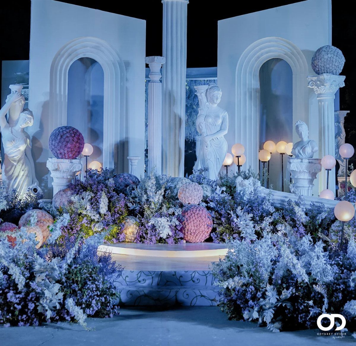 Fantasy Wedding Ideas: 9 Decor, Attire and Theme Details for 2026 9 Classical white columns and statues surrounded by blue and purple flowers and glowing orb lights.