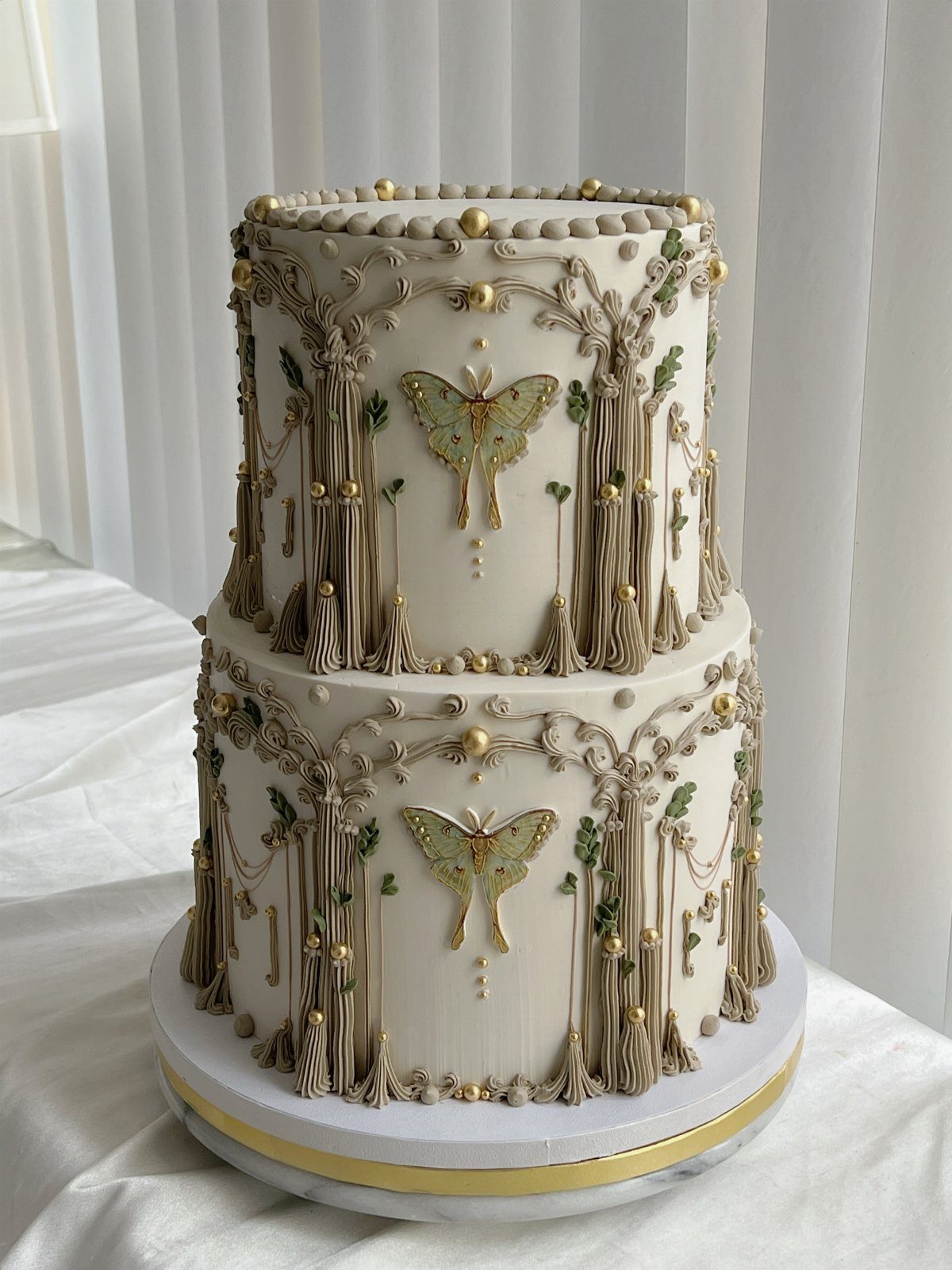 Fantasy Wedding Ideas: 9 Decor, Attire and Theme Details for 2026 3 Two-tier white wedding cake decorated with gold tree patterns and green butterfly motifs.