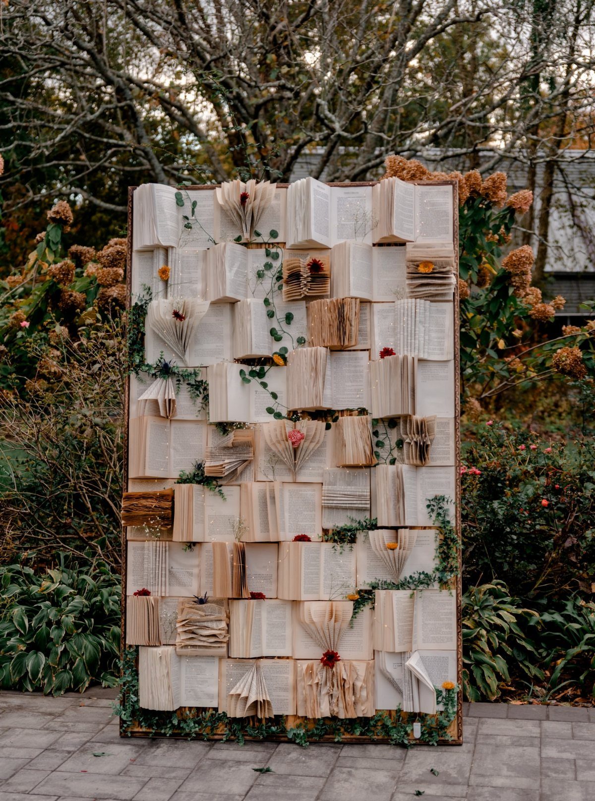 Fantasy Wedding Ideas: 9 Decor, Attire and Theme Details for 2026 8 A decorative backdrop composed of many open books adorned with small green vines and flowers.