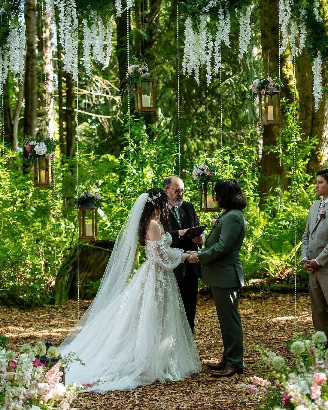 Fantasy Wedding Ideas: 9 Decor, Attire and Theme Details for 2026 2 A bride and groom exchanging vows in a forest decorated with hanging lanterns and wisteria.