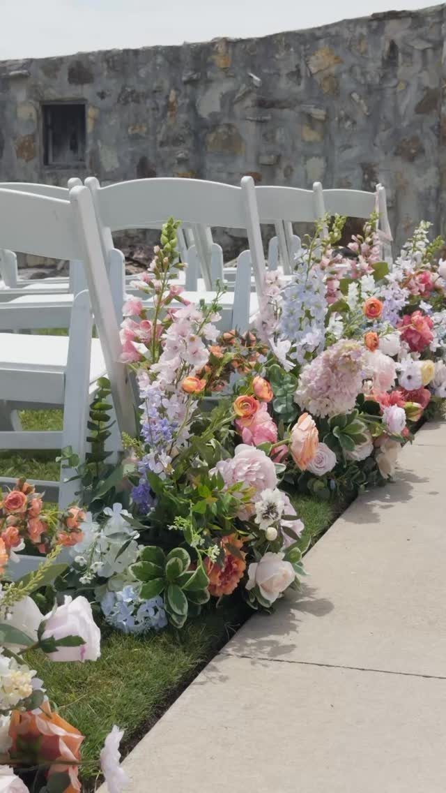 Flower Wedding Decor: 8 Ideas for Ceremony and Reception 7 White wedding chairs lined with colorful floral arrangements along a grassy walkway next to a stone wall.
