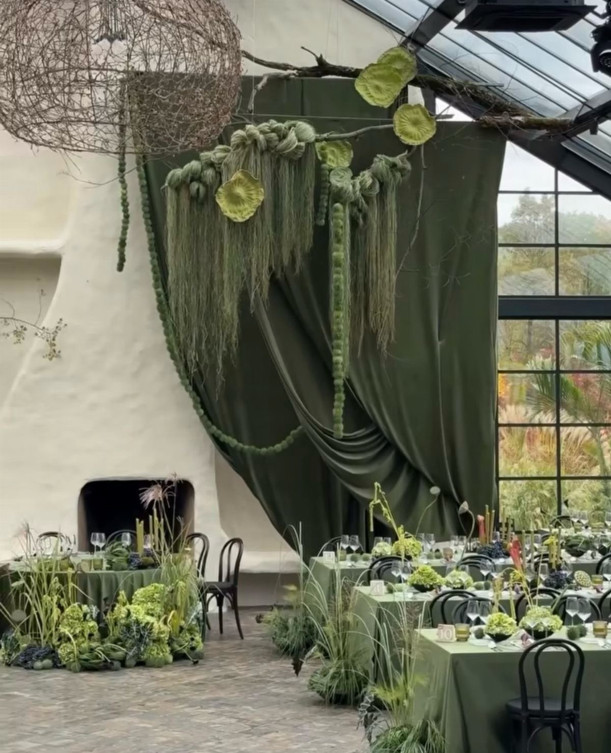 19 Forest Theme Wedding Decorations From Greenery Canopies to Candlelit Paths 2 Greenery-decorated banquet tables set in a room with large green fabric drapes and hanging plants.