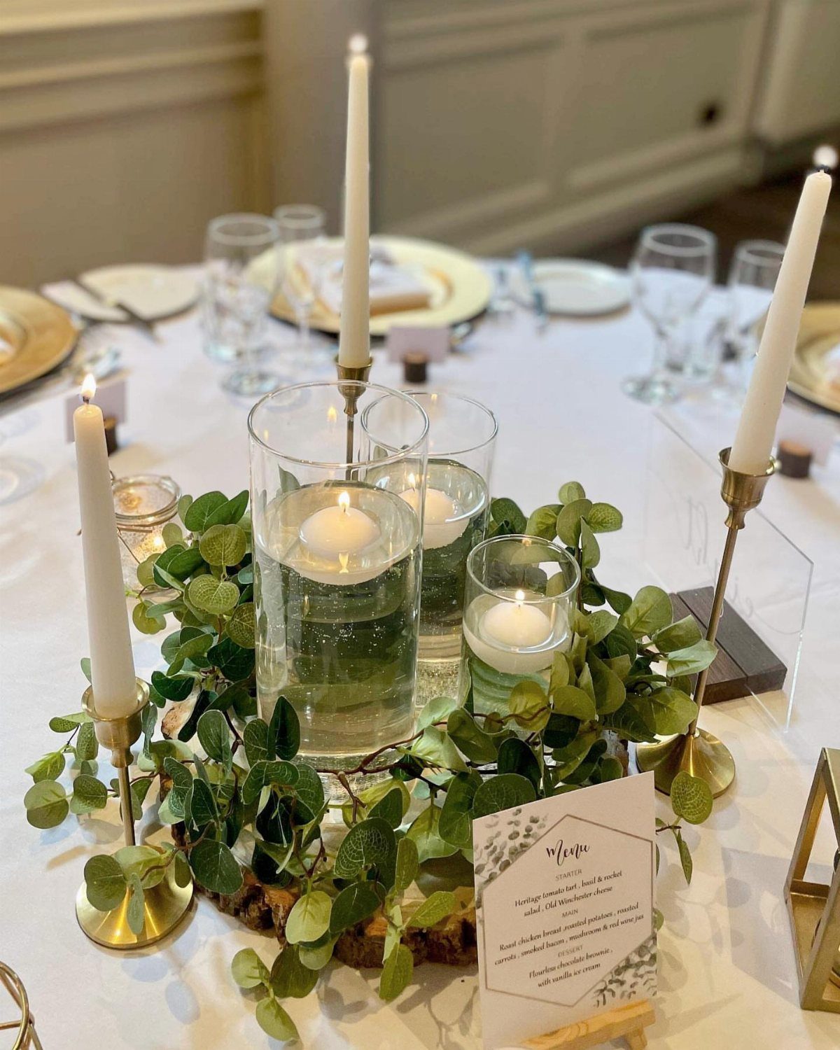 19 Forest Theme Wedding Decorations From Greenery Canopies to Candlelit Paths 6 Gold candlesticks with white taper candles and floating candles surrounded by green leaves.