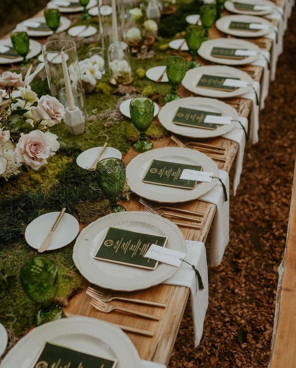 19 Forest Theme Wedding Decorations From Greenery Canopies to Candlelit Paths 8 A long wooden wedding table decorated with green moss, white plates, and elegant glassware.