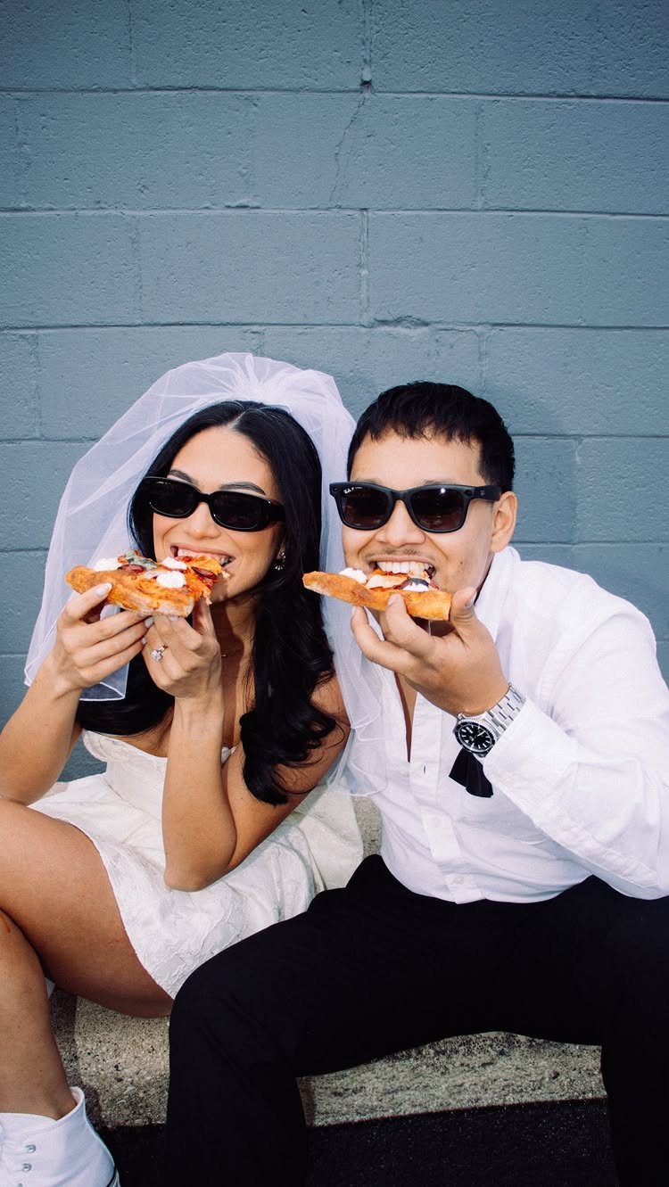 7 Funny Save the Date Ideas That Skip the Boring Part 6 Couple in wedding clothes and sunglasses eating pizza against a grey brick wall.
