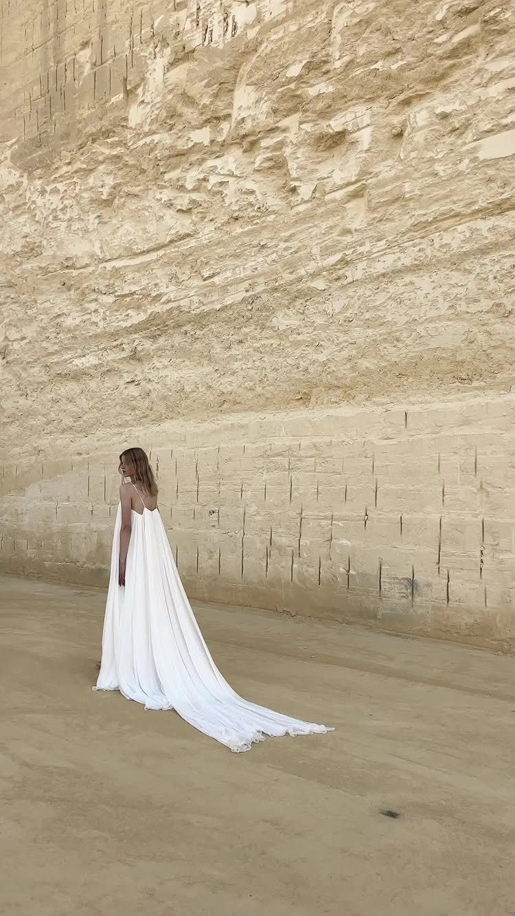 Goddess Wedding Dress Styles: 19 Ideas for 2026 17 Woman in a flowing white wedding dress standing before a tall sandy cliff.