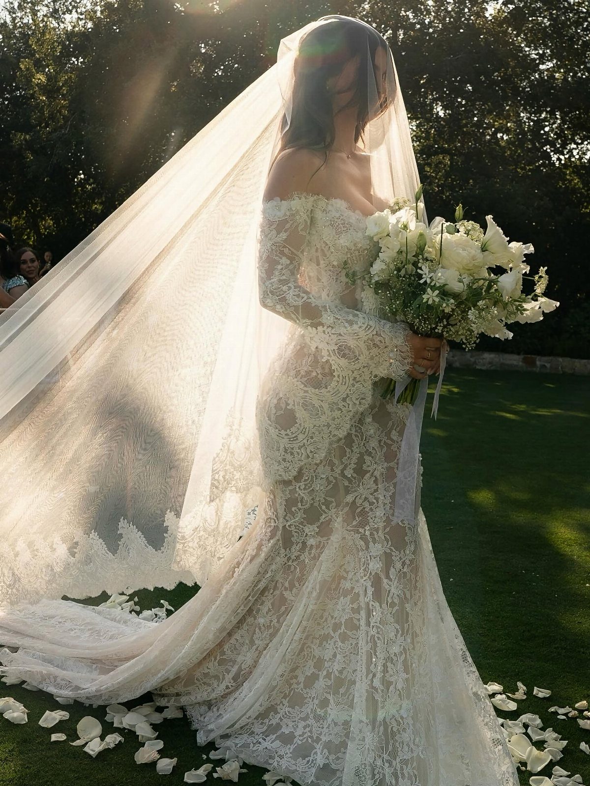 Goddess Wedding Dress Styles: 19 Ideas for 2026 5 Woman in a white lace fit-and-flare wedding gown holding white lilies on a lawn.