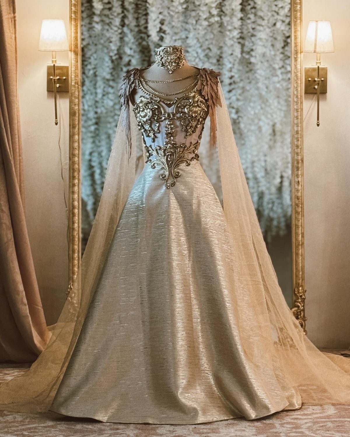 Goddess Wedding Dress Styles: 19 Ideas for 2026 13 Gold and cream goddess wedding dress with ornate bodice and sheer cape on mannequin.