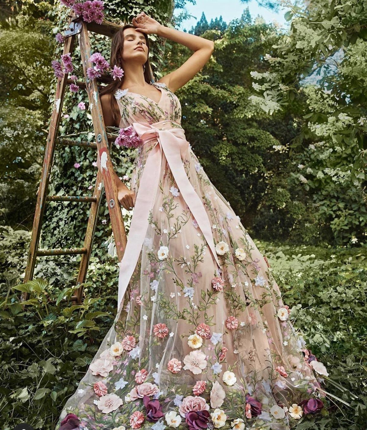 Goddess Wedding Dress Styles: 19 Ideas for 2026 4 Woman in a floral bridal gown with a pink sash posing in a garden.