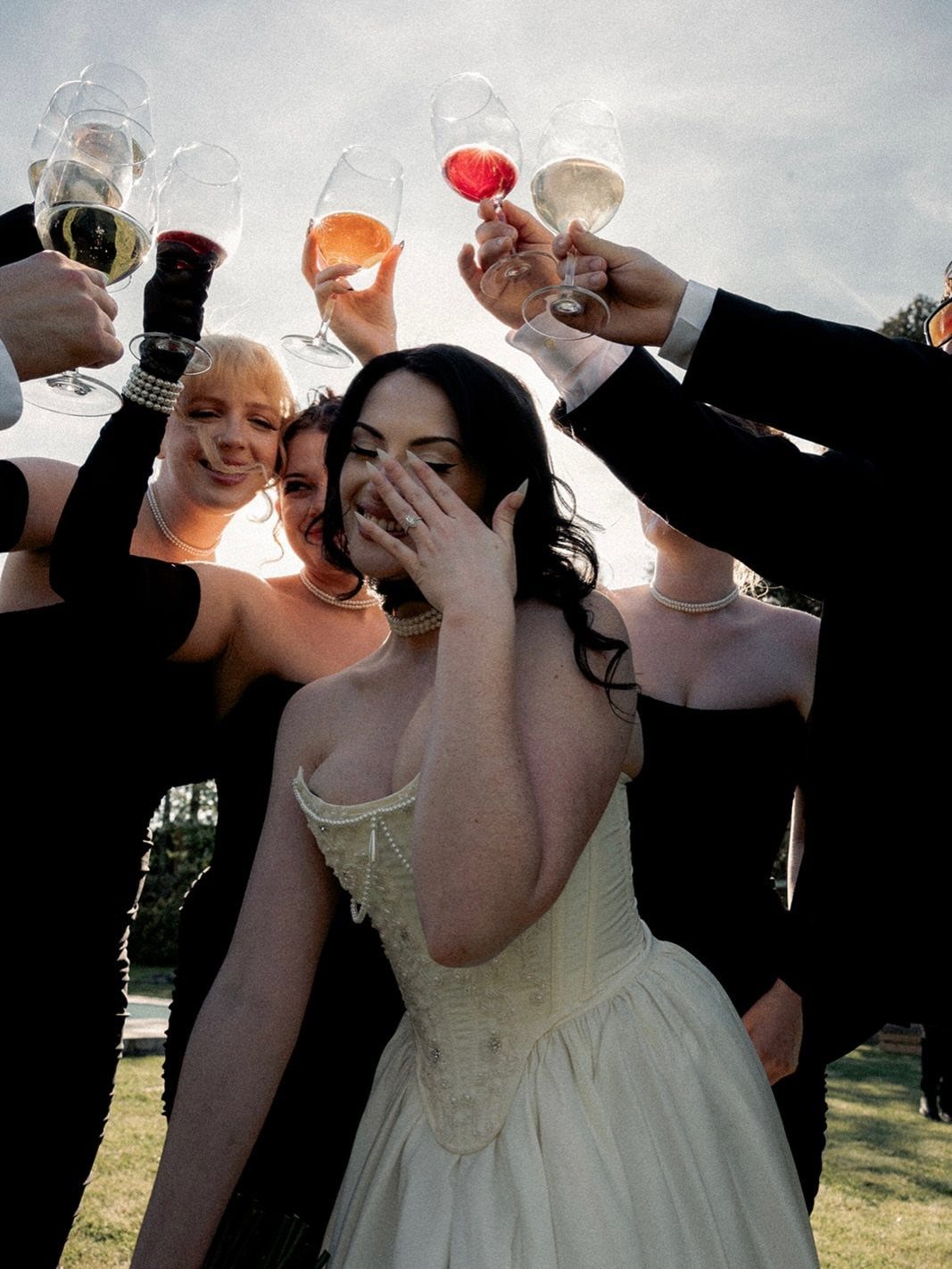 Bride in white dress and bridesmaids in black dresses raising champagne glasses in toast.