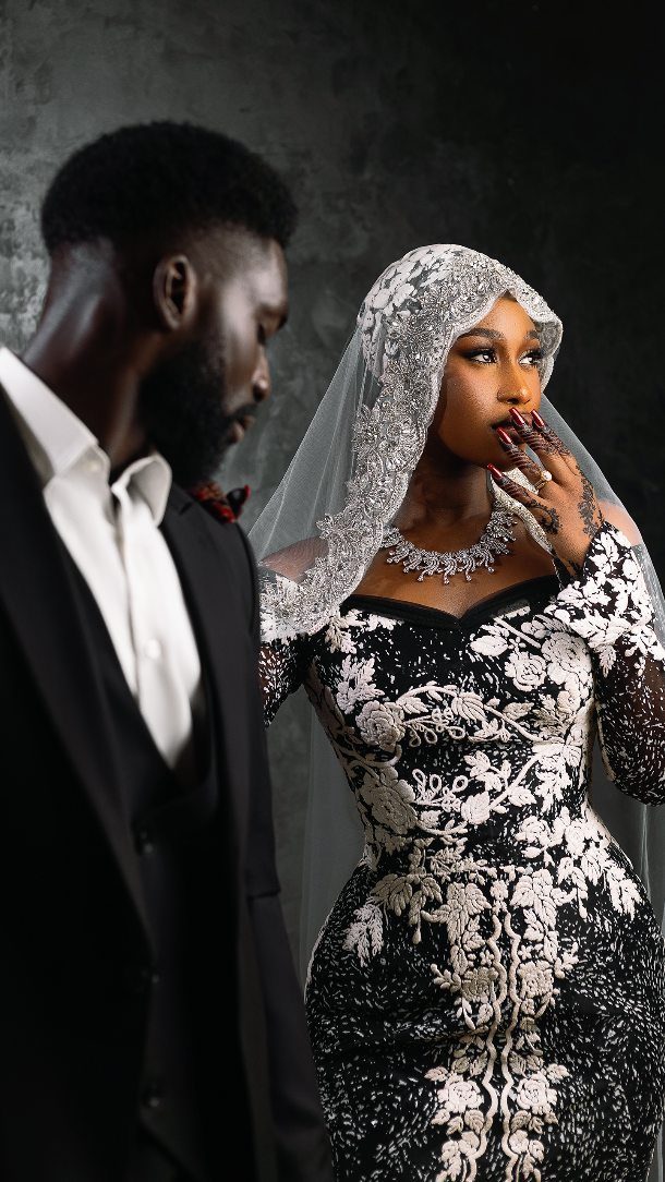 Couple in black and white wedding attire posing against a dark grey wall.