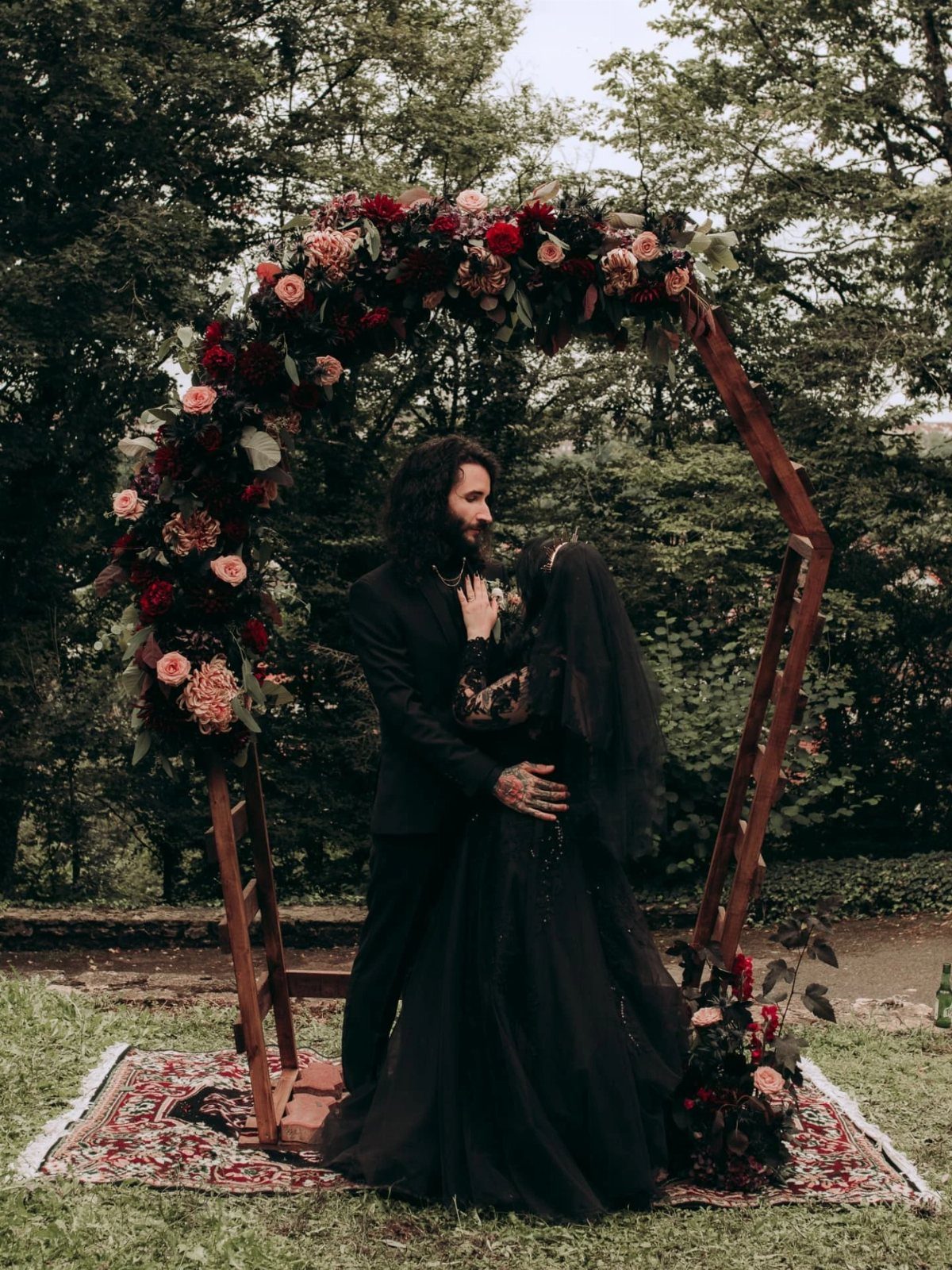 Couple in black wedding attire embracing under a floral hexagonal arch outdoors