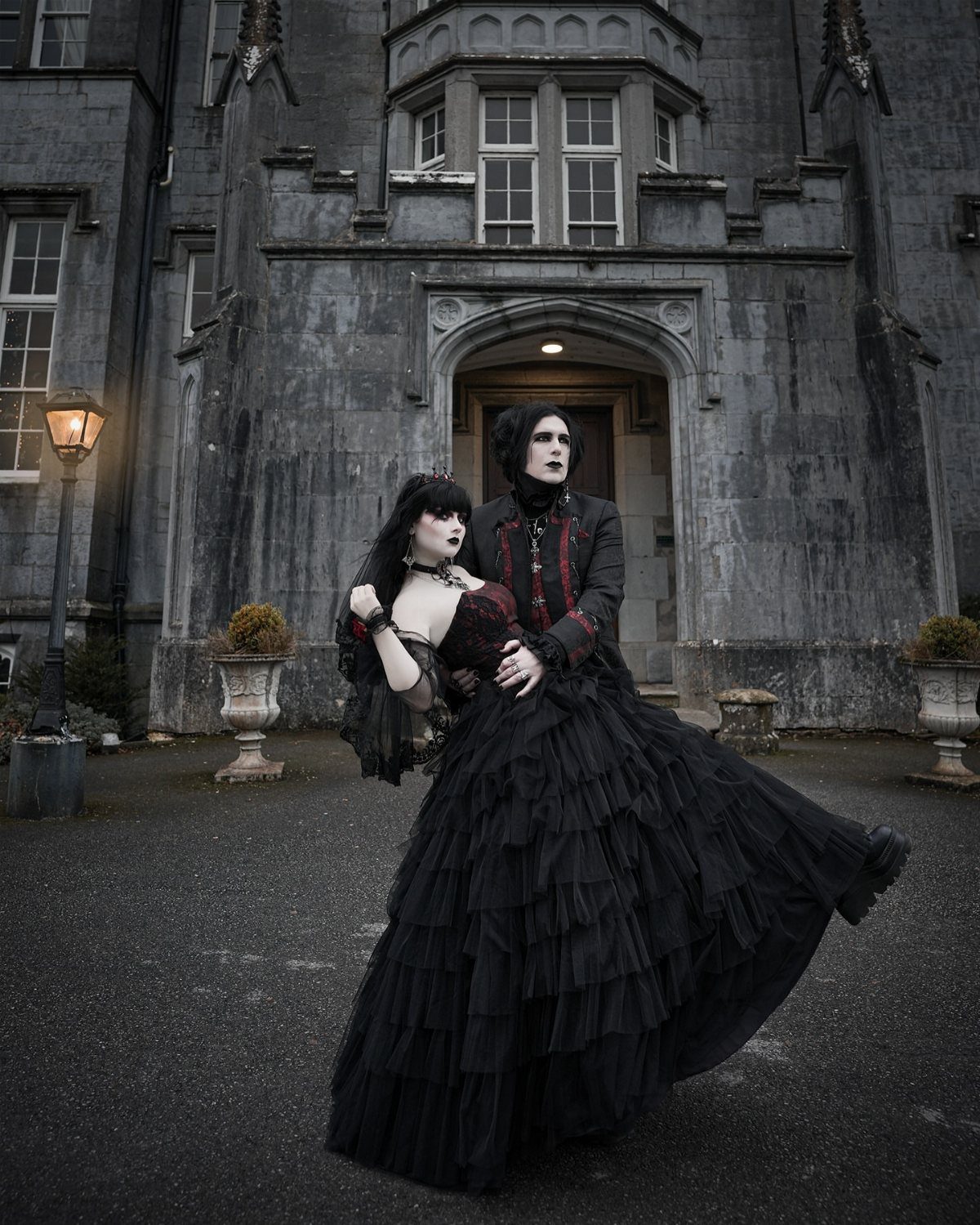 Gothic couple in black formal wear posing in front of a stone castle.
