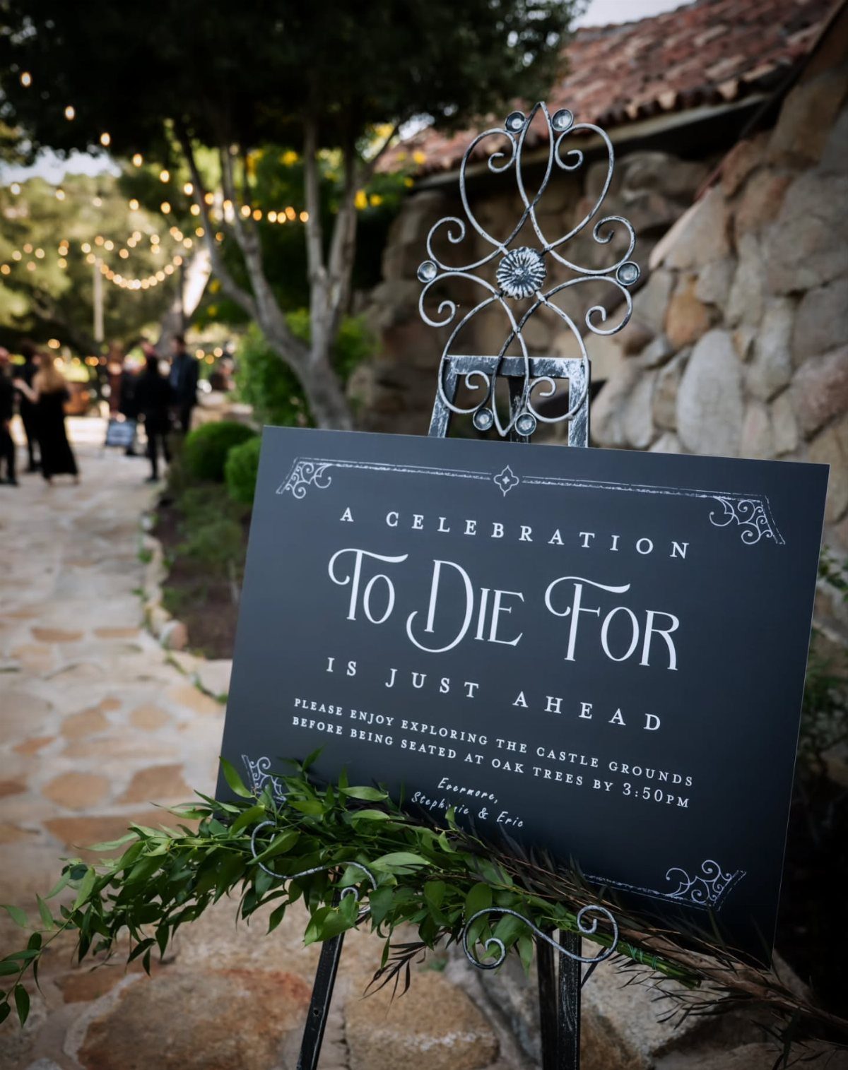 Black welcome sign with white text on a stone path with greenery.