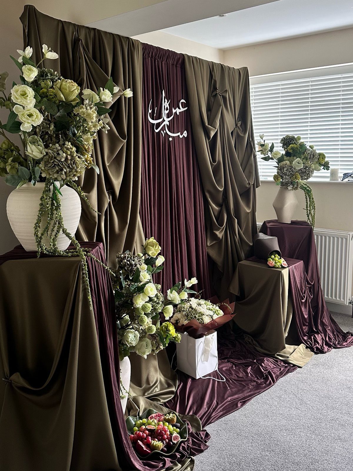 Olive green and purple draped fabric backdrop with white floral arrangements and Arabic calligraphy.